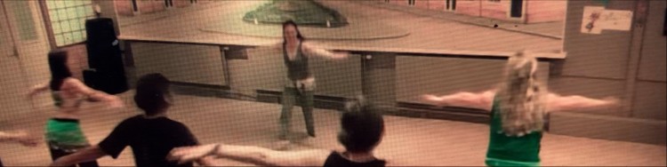 Buikdanslerares Cynthia Dorrestijn doet in spiegelbeeld een beweging voor in de danszaal tijdens een bellyrobics les van Raqs Enschede en vier dames, gezien vanaf de rugkant, imiteren de beweging.